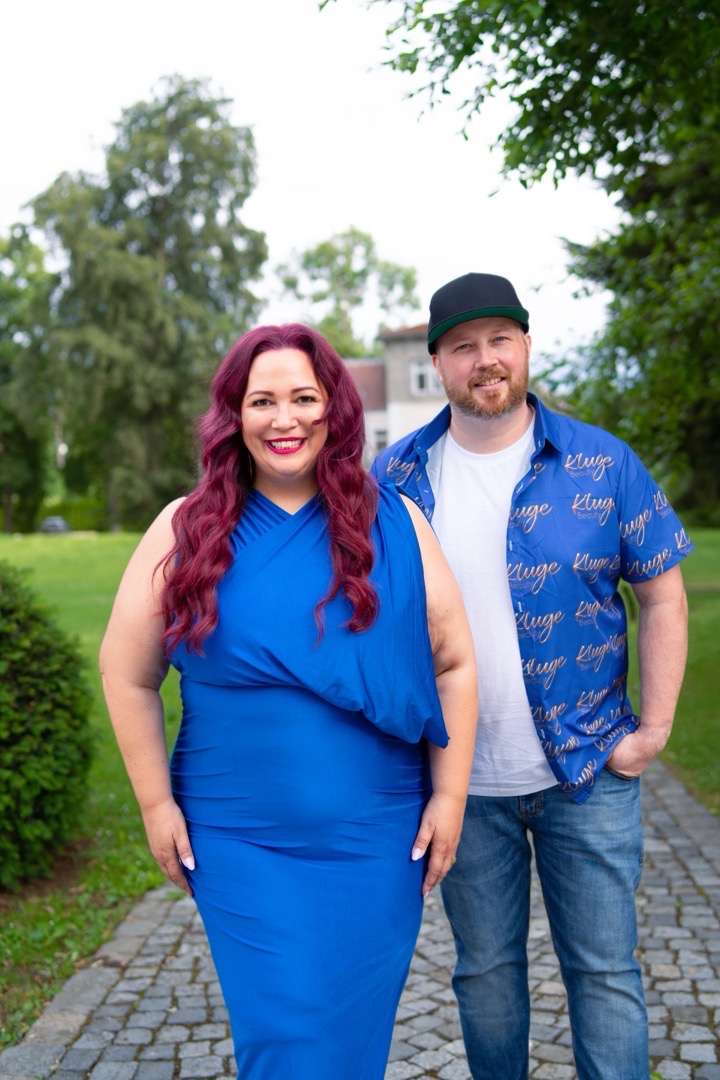 Nadine Kluge im königsblauen Kleid und ihr Mann Basti im KLUGE-Hemd stehen nebeneinander auf einem Parkweg
