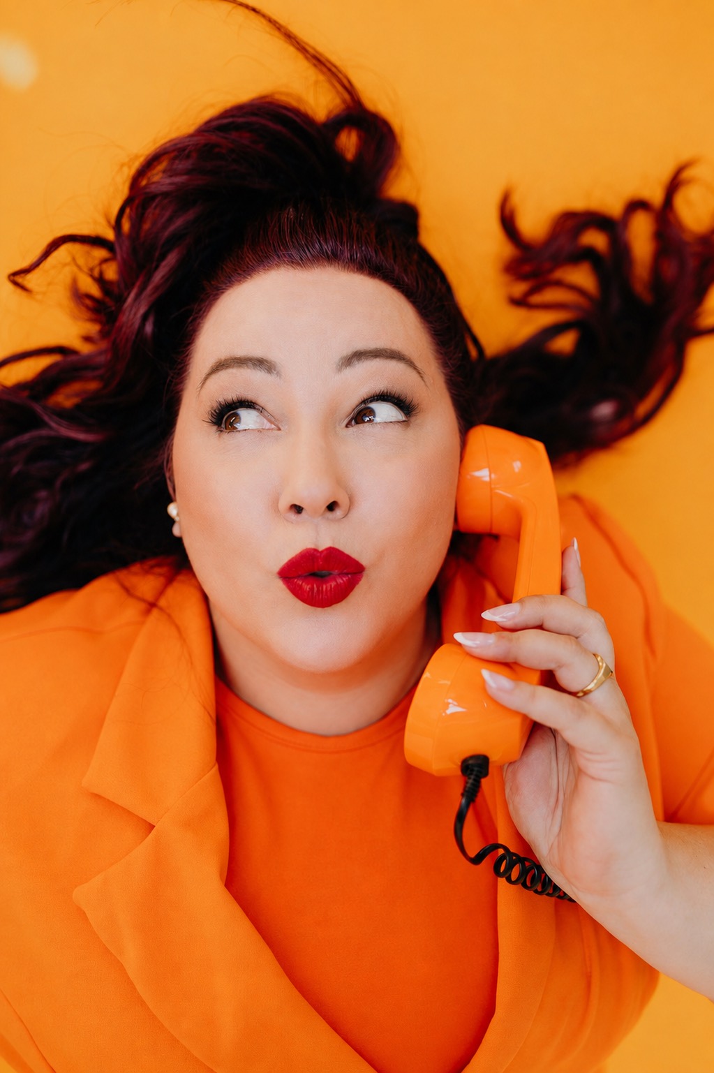 Nadine Kluge liegt auf orangefarbenem Grund, das Haar ausgebreitet, und hält einen orangefarbenen Telefonhörer ans Ohr